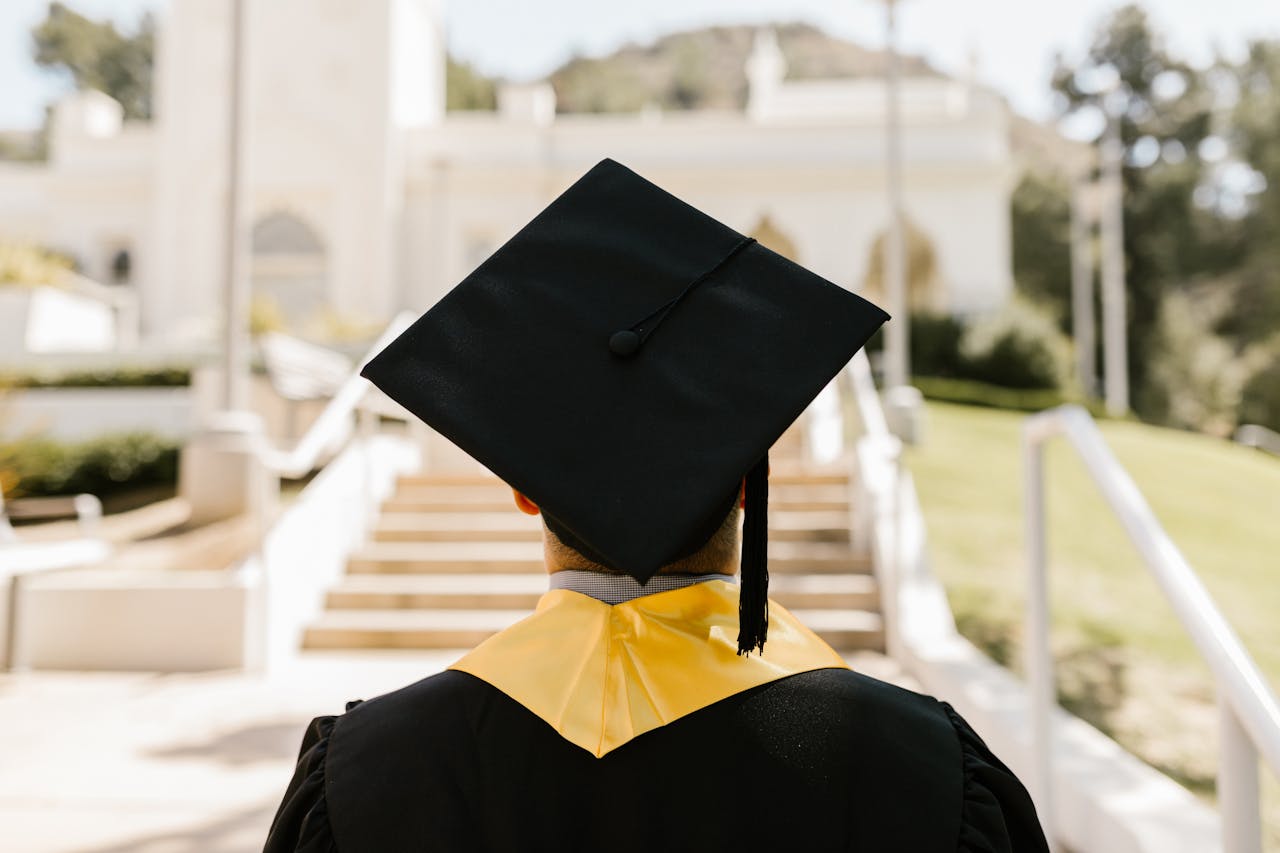 Back view of a graduate in a cap and gown standing outdoors facing a building.